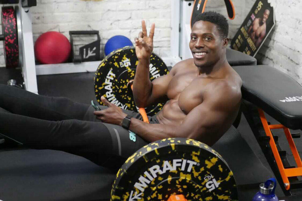 Mirafit M3 7ft 20kg Olympic Barbell Harry Aikines-Aryeetey posing while sitting underneath a Mirafit Olympic Barbell and bumper plates