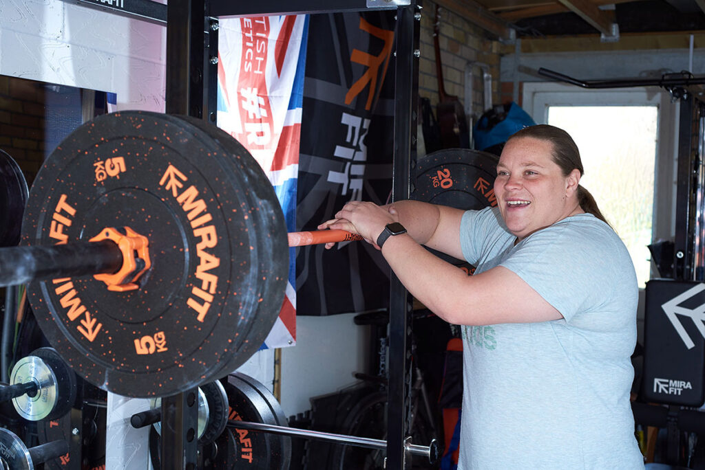 Sophie McKinna standing with a Mirafit Barbell and 5kg Weight Plates in a garage gym