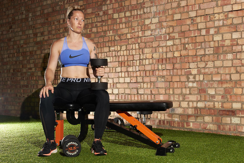 Mirafit Rubber Dumbbell Set - 12.5kg Dumbbells Woman sitting on a Mirafit weight bench while holding a Mirafit 12.5kg dumbbell in her hand